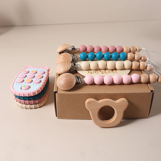Set of baby teething toys including a Pink Teether with phone buttons, wooden teething ring, and pacifier chains on a box, on a beige background.