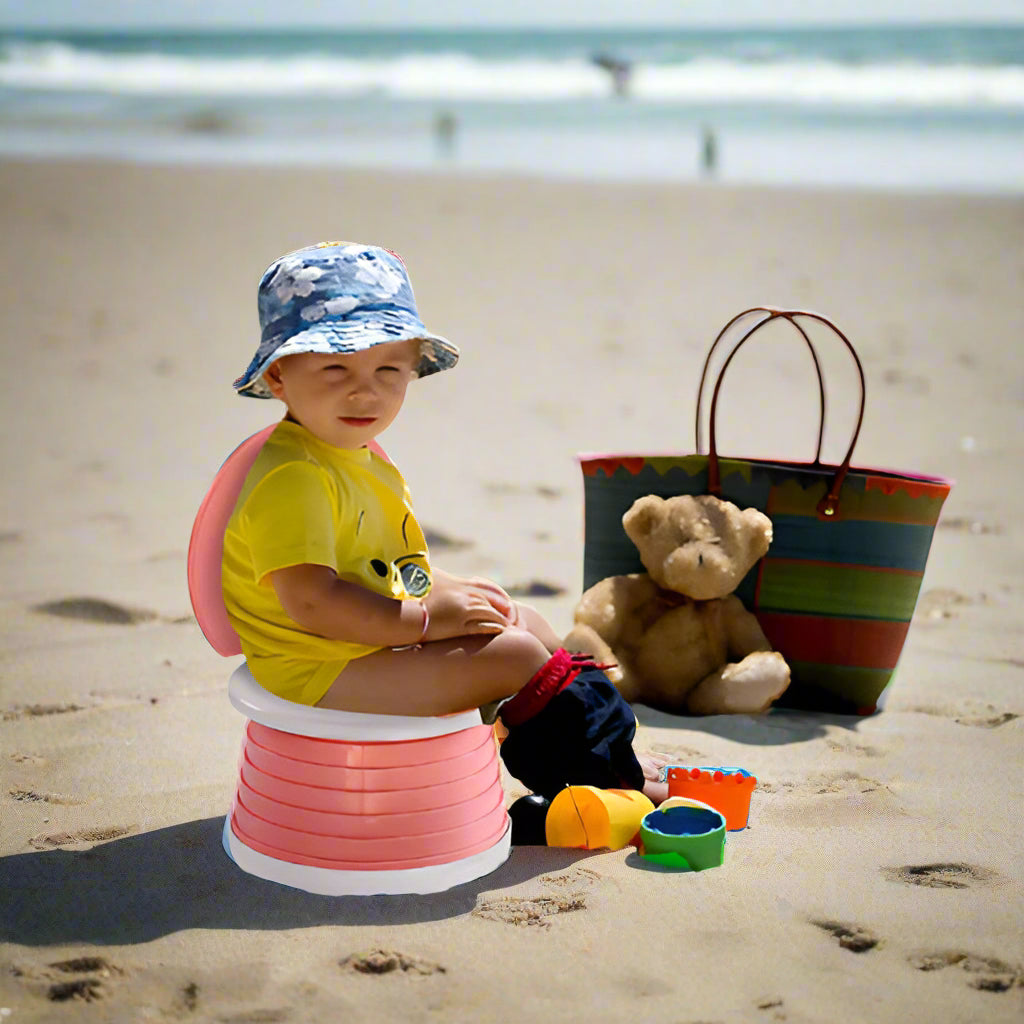 Child sitting on a pink folding Potti at the beach with toys and a teddy bear.