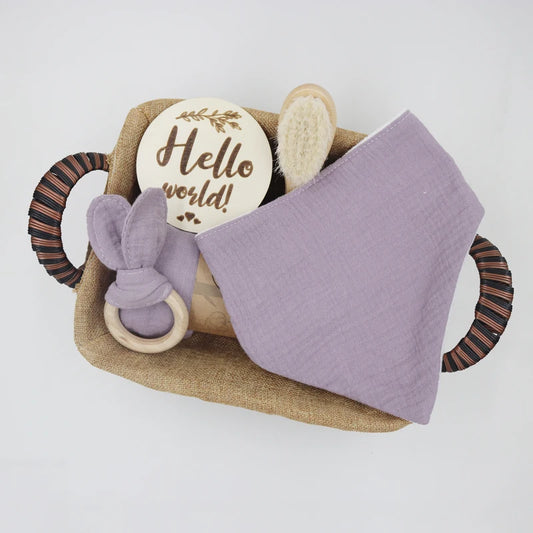 Purple Baby care set with a wooden plaque, brush, Teether and blanket in a woven basket on a white background
